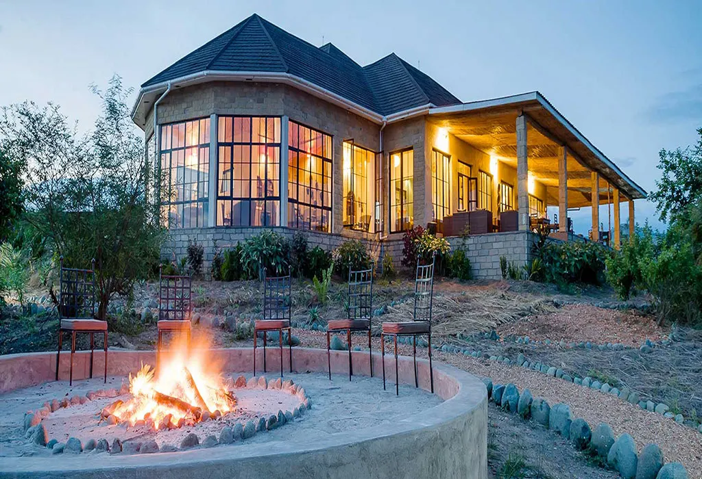 Stone Elephant Plains Lodge main building with large glass windows glowing at dusk beside a lit campfire and metal chairs.