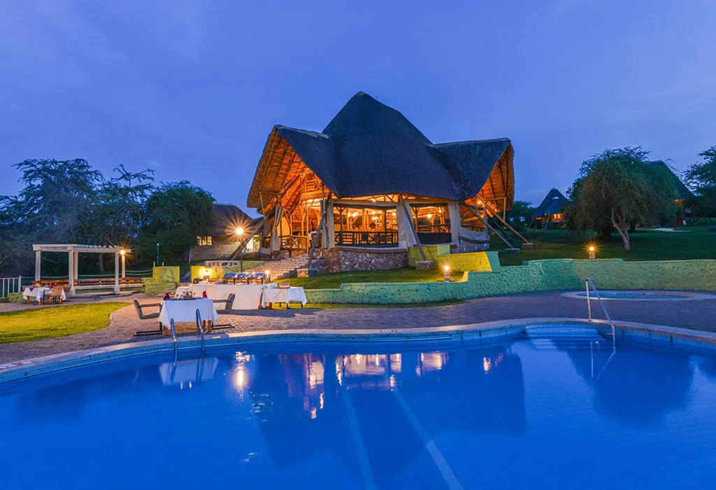 Kigambira Safari Lodge at dusk, with its illuminated thatched restaurant building beside a glowing blue swimming pool