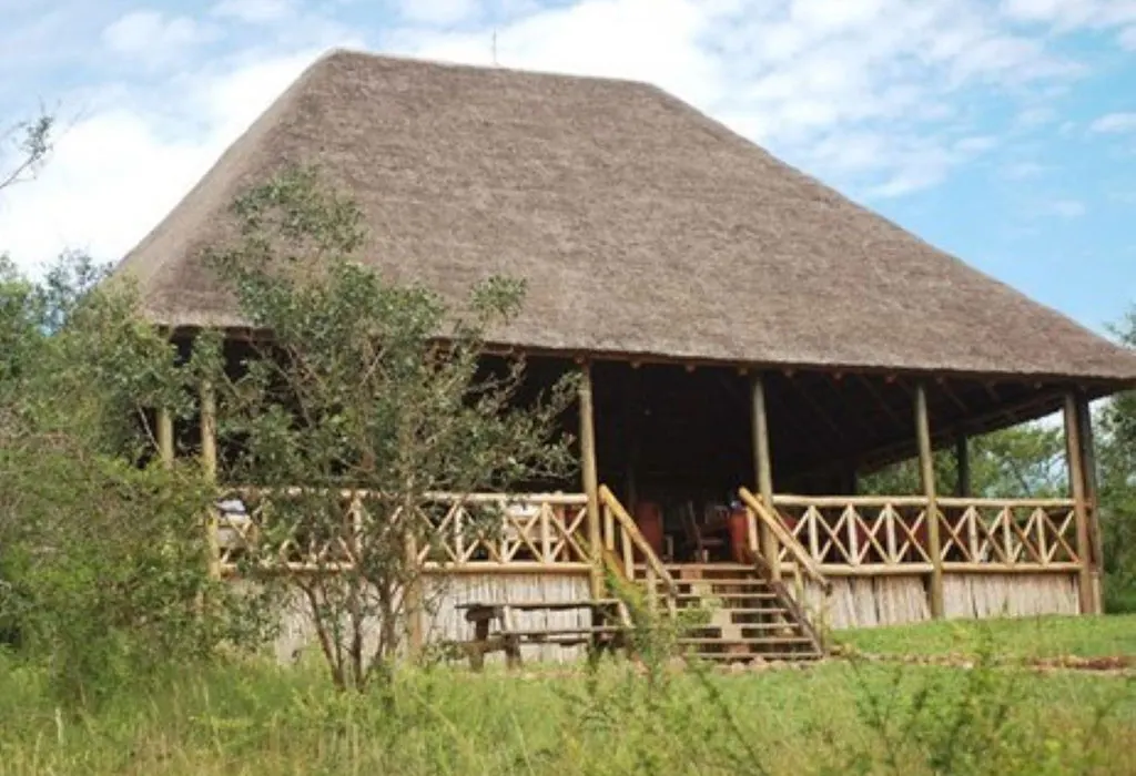 Large thatched-roof safari lodge building on stilts with a wooden veranda and railing, surrounded by green bush