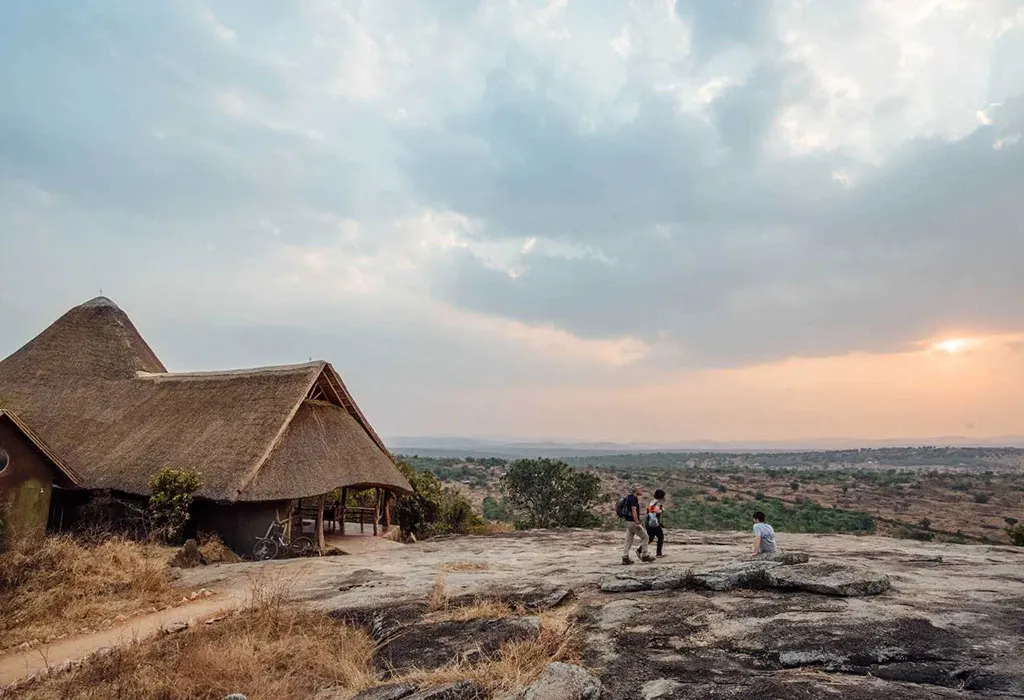 Thatched lodge perched on a granite rock outcrop at sunset, with two guests walking across the rock toward wide views of the savannah