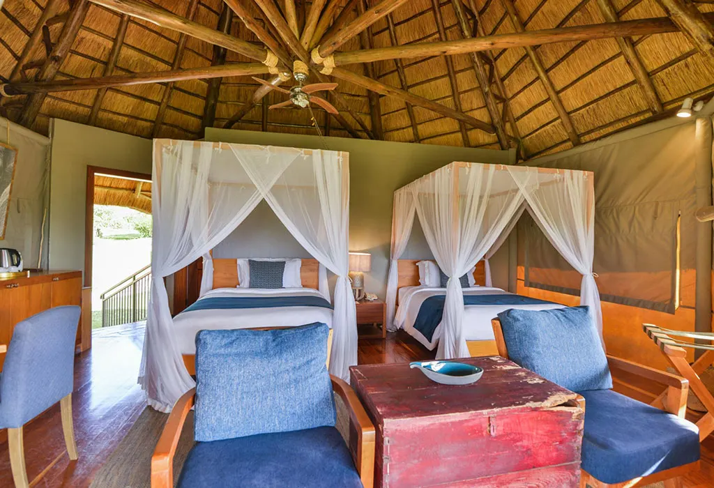 Spacious deluxe twin room with two canopy beds draped in white nets, blue armchairs and a thatched ceiling with fan