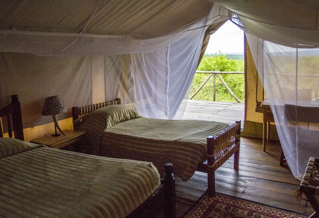 Interior of a safari tent with two twin beds under mosquito nets, opening onto a wooden deck overlooking green bush